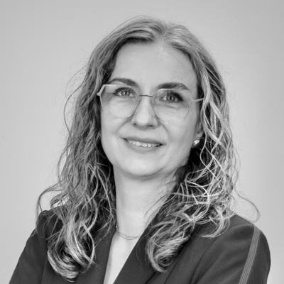 Black and white image of a Camelia, a woman with curly hair that extends beyond her shoulders and rectangular thin-framed glasses. She wears a gentle smile and a blazer.