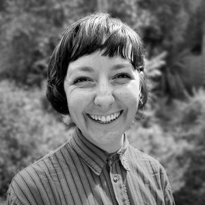 Image of young woman with a dark-haired bob and bangs. She wears a vertically-striped shirt and a big smile that squints her eyes.