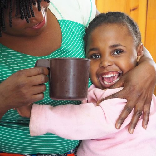 Photo of a smiling young girl in Zambia sitting on her mother's lap. The mother is holding a mug.