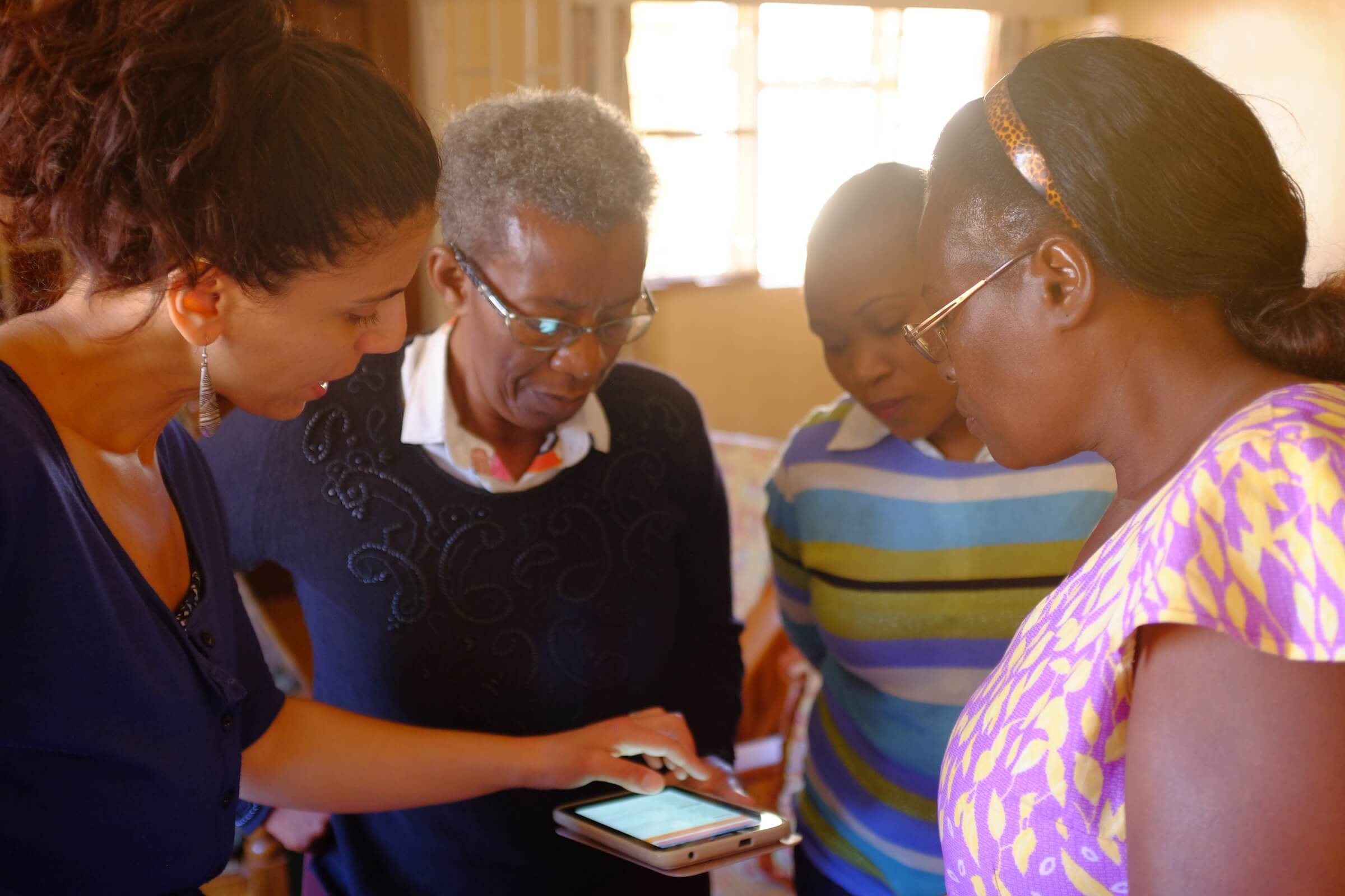 SPOON staffer Zeina Makhoul shows three women in Zambia how to use our app Count Me In, which they are looking at on a tablet.